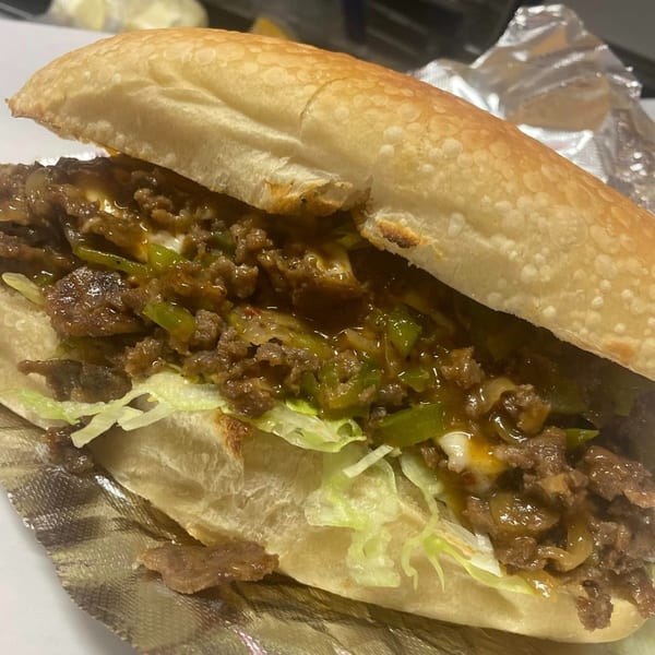 A close-up of a Lina's cheesesteak sandwich with meat, peppers, onions, and lettuce on a bun.